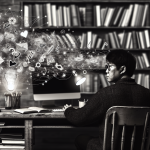 black and white image of a writer at a desk with a computer. Artistic notes, thoughts, and ideas are seen swirling around, indicating the blend of AI and human creativity